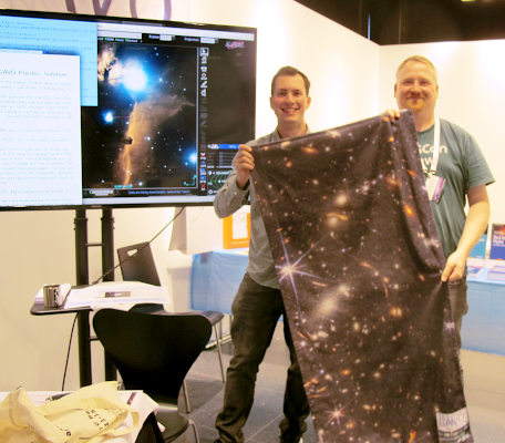 Two persons holding a large towel with an astronomical image printed on it, in the background a big screen with the Aladin VO client on it.