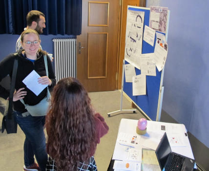 Chatting people; one is looking at a poster wall with many loose notes and a poster and is smiling.