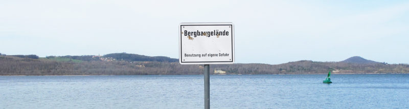 A lake with hills behind it; in it a sign saying “Bergbaugelände/ Benutzung auf eigene Gefahr”