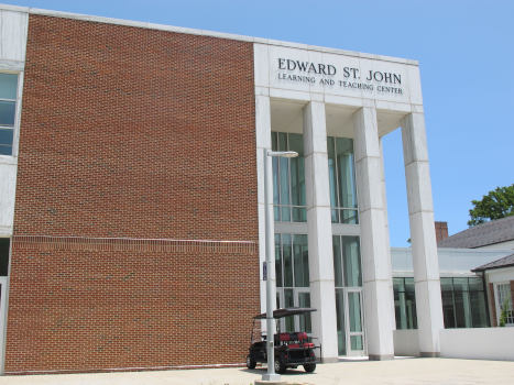 A part of a modern-ish square building, partly clinker brick, partly concrete pillars with glass behin them, holding a portico saying “Edward St. John Learning and Teaching Center“.