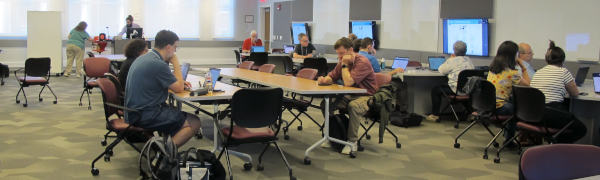 A large room with a some relatively scattered people around tables looking at various screens.  At the far end of the room, there are windows and a lectern.