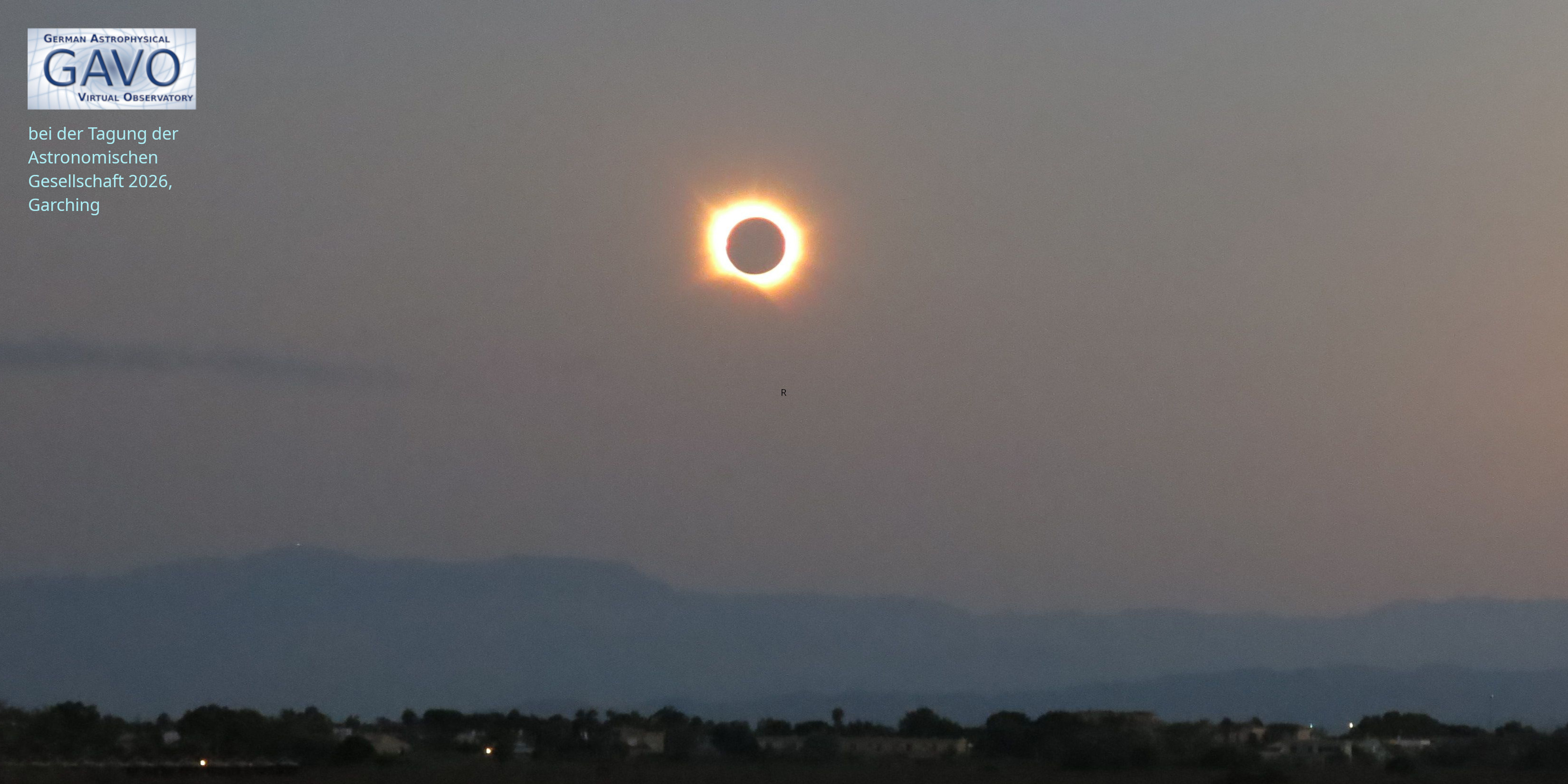 A total solar eclipse with a fairly round corona, not far above a horizon with mountains behind a plane with a few street lights.