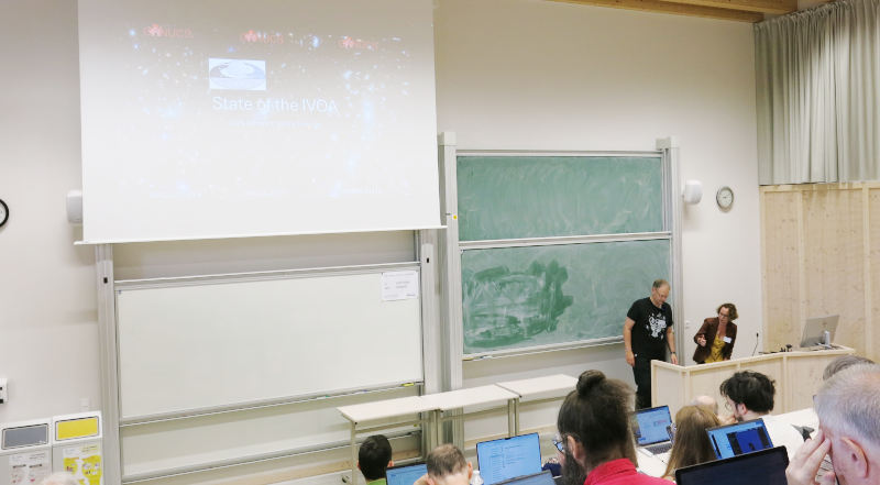 A lecture all with raised seating and a blackboard, two persons behind a wide lectern preparing for a talk.  On the wall, a slide with the title “state of the IVOA” is shown.
